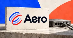 One large official Aero logo with its blue, lavender and red pinwheel and dark wordmark on an ivory stone sign beside a smaller unbranded greyscale network switch with two connected cables, against cobalt and coral panels.