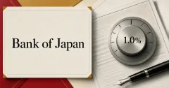 Large official Bank of Japan wordmark on an ivory policy-document plaque beside a grayscale 1.0% interest-rate dial, policy papers and a fountain pen.