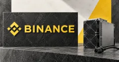 One large official yellow Binance emblem and BINANCE wordmark on a charcoal stone sign beside an unbranded greyscale ASIC mining machine with a disconnected cable, against yellow, charcoal and off-white panels.