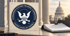 Large official navy, gold and white CFTC circular seal on an off-white stone desk sign beside an unbranded greyscale regulatory folio with blank papers and a binder clip, against a blurred Washington architectural backdrop.