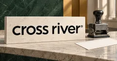 Large official black cross river wordmark on an off-white limestone banking desk sign beside an unbranded greyscale mechanical transfer stamp and blank deposit slip, against forest-green and ochre panels.
