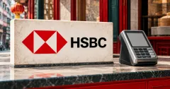 Large official red HSBC hexagon and black HSBC wordmark on an ivory merchant-counter sign beside an unbranded greyscale payment terminal with a blank display, against a Hong Kong shopfront with red framing and deep petrol trim.