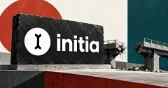 One large official white Initia icon and lowercase wordmark on a charcoal stone sign beside an unbranded greyscale broken bridge, against off-white, rust-red and deep-petrol panels.