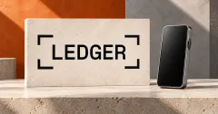 Large official black LEDGER wordmark with four corner brackets on an off-white limestone sign beside a smaller unbranded greyscale handheld touchscreen with a blank dark display, against orange and slate panels.