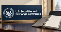 Large official white SEC seal and wordmark on a navy and gold sign beside an unbranded greyscale open ring binder with blank tabbed pages on a desk.
