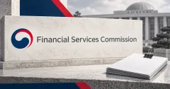 One large official navy and red Financial Services Commission emblem and English wordmark on an off-white stone sign beside unbranded greyscale securities paperwork and a metal binder clip, with navy and red panels and a blurred institutional building.