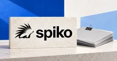 Large official black Spiko hedgehog logo and lowercase spiko wordmark on an off-white stone desk sign beside an unbranded greyscale fund folio with blank papers and a binder clip, against cobalt and pale blue panels.