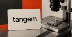 Official black Tangem wordmark on a large off-white hardware-wallet card beside an unbranded greyscale chip under a red laboratory laser beam.